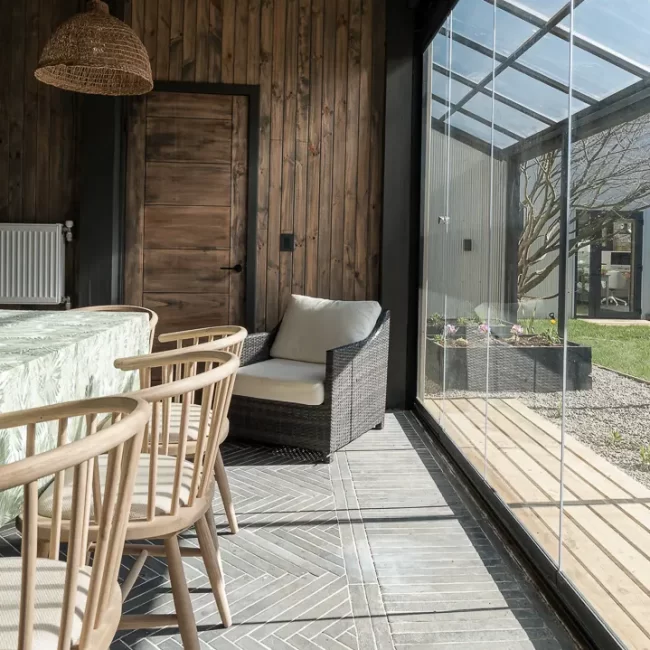 Espacio intermedio tipo sunroom con grandes ventanales y estructura de madera carbonizada, diseño bioclimático para el sur de Chile.