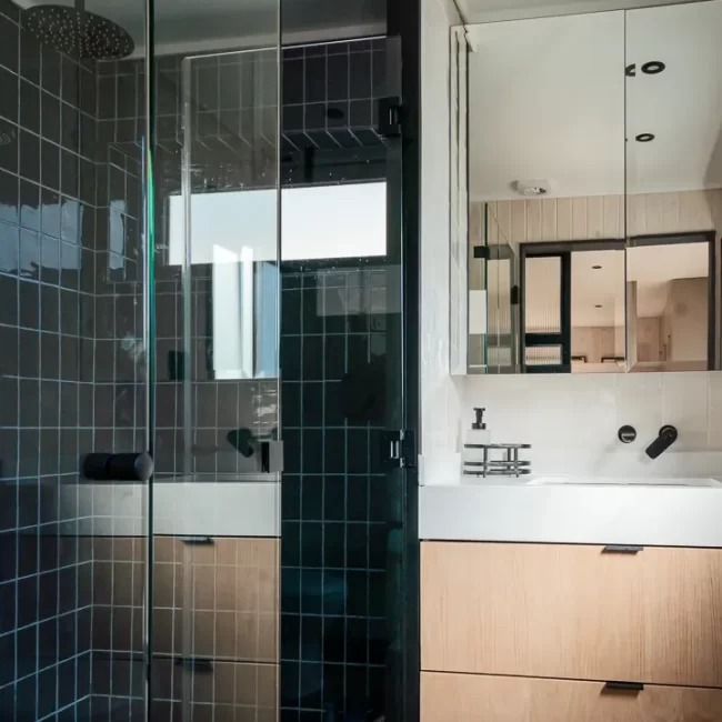 Detalle de baño remodelado con ducha walk-in, mampara de vidrio y grifería negra mate, acabados modernos en proyecto residencial.